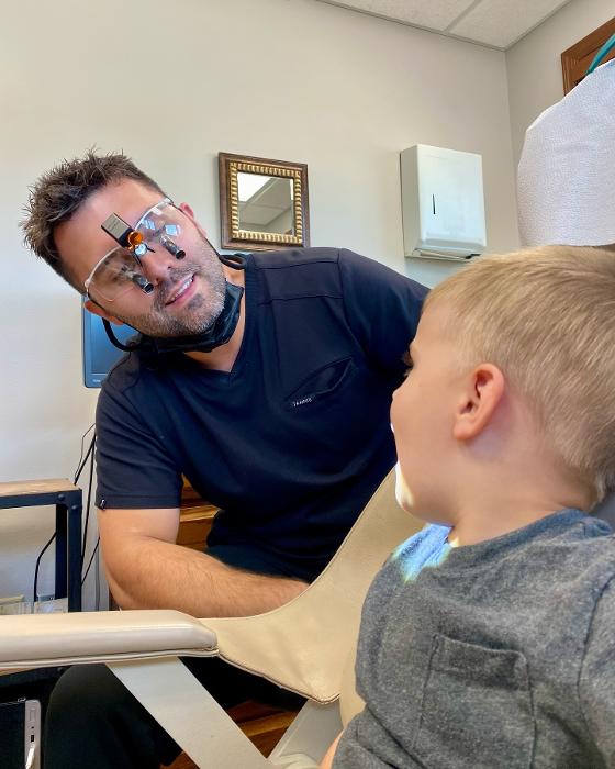 A young boy sits in a dental chair while a dental professional in scrubs speaks to him, illustrating a friendly and welcoming children's dentistry environment.
