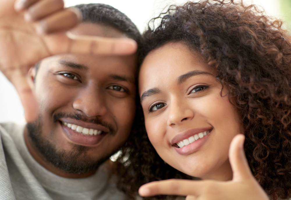 Happy couple smiling with bright teeth, showcasing the results of a smile makeover, while framing their faces playfully with their hands
