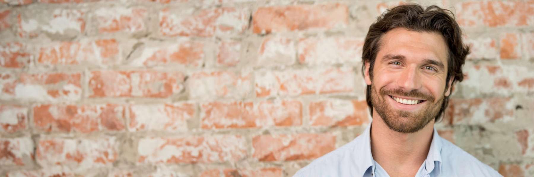 Confident man with a bright, flawless smile standing against a brick wall, highlighting the benefits of a smile makeover